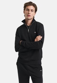 Black sweatshirt with a quarter-zip collar and ribbed cuffs. Features a small logo on the left chest and a soft, smooth texture.