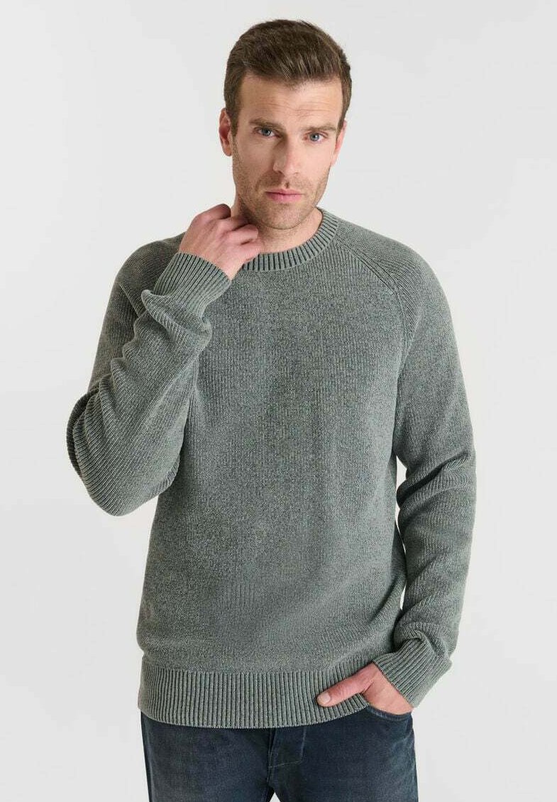 Mann mit kurzen braunen Haaren, der einen grünen Strickpullover und dunkle Jeans trägt, steht mit einer Hand in der Tasche und der anderen Hand am Hals.