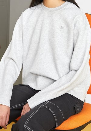 Personne assise sur une chaise orange portant un sweat-shirt Adidas gris clair et un pantalon noir avec des coutures blanches.