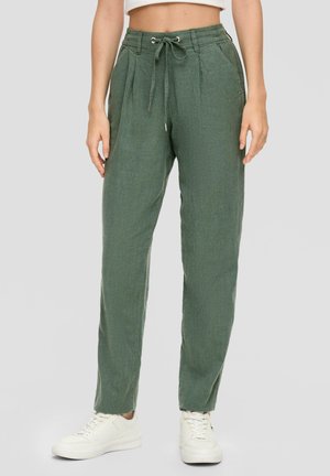 Femme portant un pantalon vert à cordon et des baskets blanches, debout devant un fond clair uni.