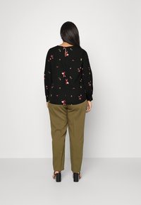 Kvinde med langt mørkt hår iført en sort blomsterbluse, olivengrønne bukser og sorte højhælede sandaler, der står med ryggen til.