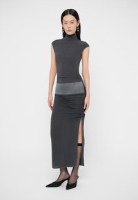 Szürke, testhez simuló ruha magas nyakkal, ráncolt részletekkel, oldalsó hasítékkal és kontrasztos, fényes derékpánttal. Fekete magassarkú cipővel párosítva.