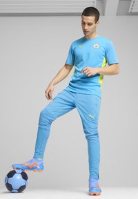 Gaiši zils futbola tērps ar dzelteniem akcentiem, ar reljefu rakstu. Modelis stāv uz melna un gaiši zila futbola.
