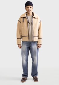 Chaqueta de piel sintética beige con detalles en marrón oscuro, cremalleras y un cuello de chal, combinada con unos vaqueros desgastados de color azul claro y zapatos marrones.