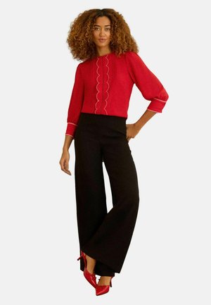 Femme aux cheveux bouclés portant un chemisier rouge avec un bord festonné, un pantalon noir à jambes larges et des chaussures rouges à talons hauts, debout devant un fond uni.