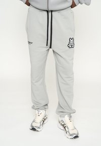 Pantalones deportivos de color gris claro con un cordón negro, que presentan un logo en el muslo izquierdo y una textura suave. Combinados con zapatillas blancas y plateadas.