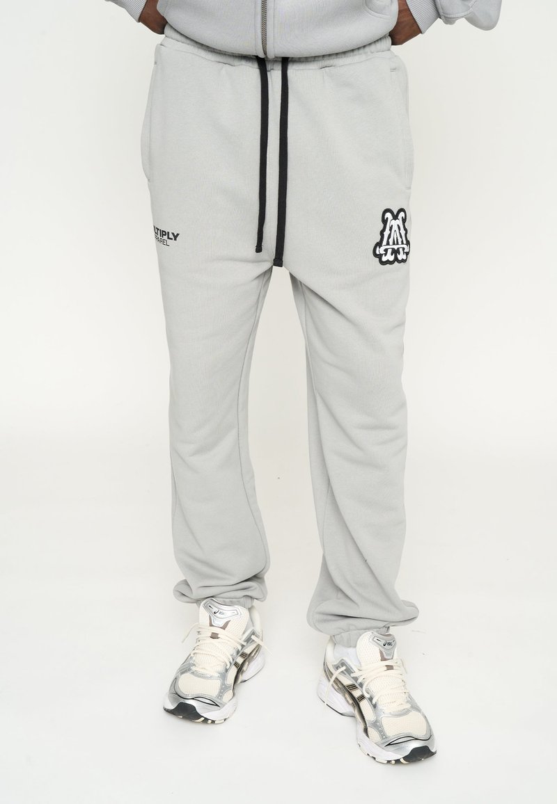 Pantalones deportivos de color gris claro con un cordón negro, que presentan un logo en el muslo izquierdo y una textura suave. Combinados con zapatillas blancas y plateadas.
