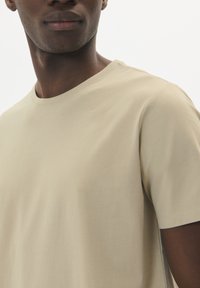 Tricou scurt cu mânecă, de culoare bej deschis, fabricat dintr-un material moale din bumbac, cu o textură netedă și cusături minime.