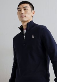 Tengerészkék pulóver negyed cipzárral és bordázott gallérral. Jellemzője egy apró, hímzett minta a mellrészen és texturált kötött anyag.