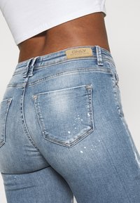 Jeans de mezclilla azul claro con detalles desgastados, costuras en contraste, dos bolsillos en la parte trasera y una etiqueta de cuero de marca en la cintura.