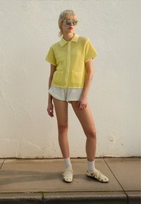 Gul nättröja med korta ärmar, krage och två fickor, kombinerad med vita texturerade shorts och dekorativa ljusa skor.