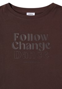 Chemise marron foncé avec texte en relief "Follow Change Dance" et texte plus petit "Live your own life, pursue your own way" sous l'encolure.