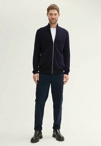 Mornarsko modra jopica z zadrgo v pletenem materialu, z visokim ovratnikom in dolgimi rokavi, kombinirana s temnimi hlačami in črnim obutvim.