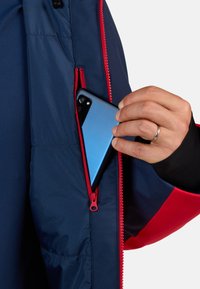 Giubbotto blu navy con accenti rossi, dotato di tasca con zip. Una mano sta inserendo uno smartphone blu nella tasca.