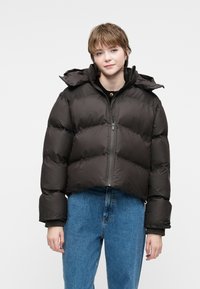 Chaqueta tipo plumón en marrón oscuro con un diseño cropped, puños acanalados y cierre frontal con cremallera. Capucha presente, con textura suave y brillante.
