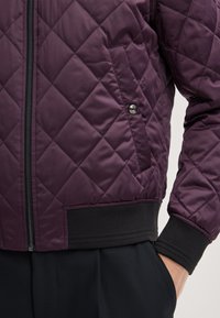 Gequilted bomberjack in donkerpaars nylon met zwarte ribgebreide boorden en tailleband, voorzien van een ritssluiting en zijzak.