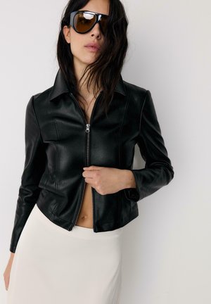 Femme portant de grandes lunettes de soleil noires, une veste en cuir noire partiellement zippée et une jupe blanche, debout devant un fond clair uni.