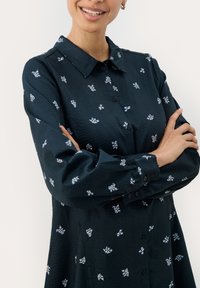 Chemise button-up vert foncé avec un tissu texturé, ornée de petits motifs floraux blancs et bleus et d'un col classique. Manches longues.