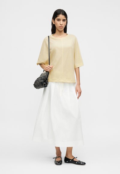Ανοιχτό κίτρινο, κοντομάνικο τοπ με στρογγυλή λαιμόκοψη· συνδυασμένο με λευκή, μακριά φούστα. Μαύρη τσάντα χειρός με πλεκτή υφή και μεταλλικά στοιχεία.