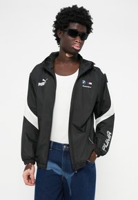 Jachetă neagră tip windbreaker cu accente albe, logo Puma și branding BMW Motorsport. Material ușor, cu închidere prin fermoar și manșete elastice.