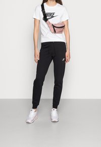 Rosa midjeväska med svart dragkedja, svarta sweatpants med logotyp, vit Nike t-shirt, vita och rosa sneakers. Neutral bakgrund.