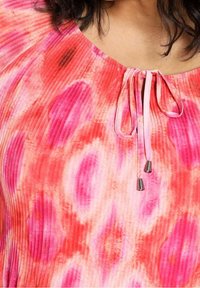 Primo piano di una blusa tie-dye rosa e rossa con tessuto plissettato e un collo con nodo caratterizzato da estremità in metallo argentato.