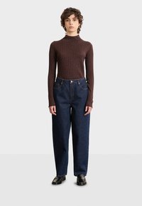Brauner gerippter Rollkragenpullover, kombiniert mit lockeren dunkelblauen Jeans, die über Fronttaschen und einen geraden Beinschnitt verfügen. Schwarze Schuhe vervollständigen das Outfit.