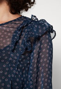 Blusa ligera de color azul marino con delicado patrón floral, detalles con volantes en los hombros y textura de tela ligera.