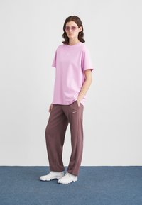 Naine, kellel on seljas roosa tooni prillid, lahti istuv roosa Nike t-särk, pruunid Nike püksid ja valged tossud, seisab sinisel vaibal.