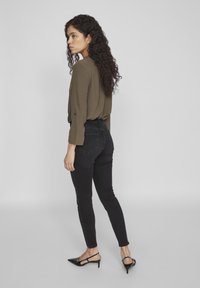 Top de manga larga verde oliva, vaqueros skinny negros y tacones de slingback negros con punta. Cabello rizado, posición de pie, mostrado de perfil.
