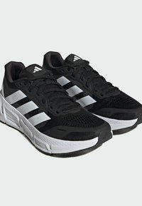 Mustat urheilukengät, joissa on verkkopäällinen ja valkoiset yksityiskohdat; varustettu kolmella valkoisella raidalla, litteillä nauhoilla ja tekstuurilla, joka takaa hyvän pidon.