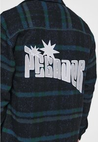 Czarna i zielona flanelowa koszula w kratę z białym haftowanym napisem "PEGADOR" i grafikami gwiazd na plecach, wykonana z teksturowanego materiału.