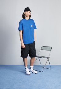 Blauwe katoenen t-shirt met een borstzak, zwarte shorts, witte sneakers met blauwe accenten, en een zwarte beanie, naast een transparante opvouwstoel.