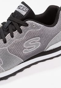 Grå atletisk sneaker med en texturerad mesh-övre, svarta accenter, snören och en vit sula. Inkluderar en framträdande logotyp på sidan.
