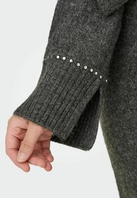 Grå stickad tröja med muddstruktur på ärmen och silverfärgade nitar längs kanten. Har en avslappnad passform och en lätt öppning vid handleden.