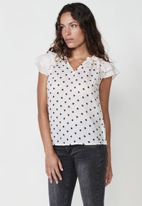 Blusa bianca con pois neri, maniche corte arricciate e scollo a V. Il tessuto appare leggero e strutturato. Abbinata a jeans scuri.