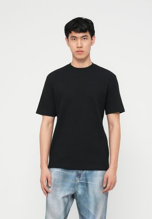 Joven con cabello negro corto, vestido con una camiseta negra de manga corta lisa y jeans azul claro, de pie contra un fondo blanco.