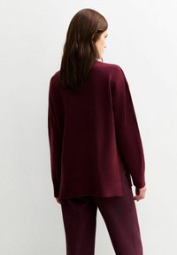 Burgundy stickad tröja med avslappnad passform, sänkt axelparti och ribbat fåll. Mjuk textur, långa ärmar och sidofickor i botten.