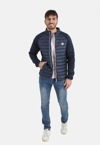 Doudoune marine avec un devant matelassé et des manches unies, dotée d'une capuche, d'une fermeture éclair et d'accents rouges. Associée à un jean bleu et des baskets blanches.