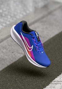 Modrá sportovní bota s texturovaným svrškem, zdobená růžovými a oranžovými akcenty. Vybavená bílým logem a polstrovanou bílou podrážkou.