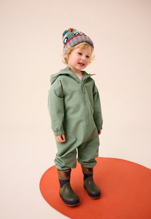 Jeune enfant portant une combinaison de pluie verte à capuche, un bonnet tricoté coloré et des bottes de pluie vertes, debout sur un tapis ovale orange.