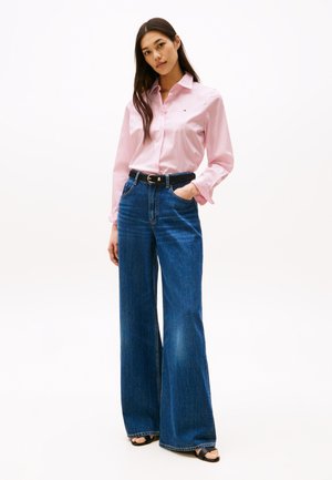En lyserød skjorte med krave parret med højtaljede, bredbenede blå jeans. Et sort bælte med en sølvspænde fremhæver outfittet. Glat stofstruktur.