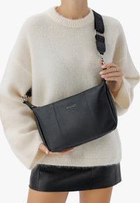 Borsa a spalla in pelle nera con texture effetto granito, hardware in argento e tracolla regolabile con motivo. Il modello indossa un maglione crema.
