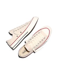 Para beżowych niskich trampek Converse z białymi gumowymi podeszwami, czerwono-czarnymi paskami i białymi sznurowadłami na białym tle.