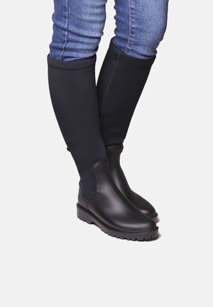 Stivali neri impermeabili al ginocchio con finitura liscia, materiale in gomma e suole rinforzate in gomma, abbinati a jeans in denim blu.
