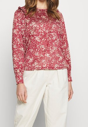 Haut rouge à manches longues avec un motif floral en crème et rose, présentant un col rond et des détails froncés aux épaules. Accompagné d'un pantalon blanc.