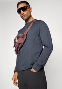 Navy long-sleeve shirt with a round neckline, featuring a front pocket. A brown crossbody bag with textured fabric is worn across the chest.