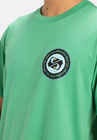 Camiseta de algodón verde con un logo redondo en negro y azul en el centro, con texto alrededor del perímetro y un diseño clásico de cuello redondo.