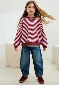Sweatshirt surdimensionné rayé rouge et rose avec des poignets côtelés, associé à un jean en denim bleu ample et des baskets bourgogne. Coiffé avec les cheveux longs.