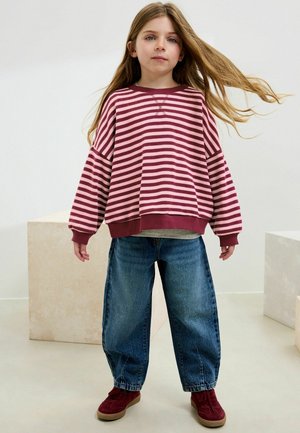 Rood en roze gestreepte oversized sweatshirt met ribbels aan de mouwen, gecombineerd met losse blauwe denim jeans en bordeaux sneakers. Gestyled met lang haar.